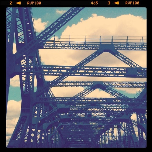 A photo of the metal structure that the Story Bridge is made out of. The section of bridge in the photo is only an abstract, and you can see the blue sky spotted with clouds through the scaffolding.