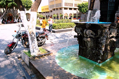Motorcycle and couple with child + trailing spray of the brilliant turquoise classical Corinthian capitol stone water fountain, windy Central square, Guadalajara, Jalisco, Mexico by Wonderlane