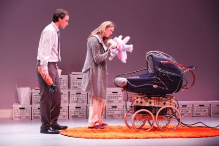 On-stage scene from the play. A man and woman stand looking into a pram, the woman with a many-limbed plush toy. The pram has a wild series of tubes and wires snaking out of it.