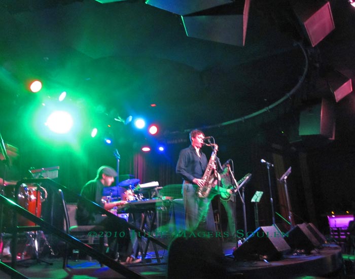 A sax player moves trailing green streamers on the stage of a small night club.