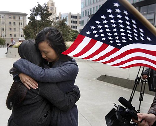 two women embrace fiercely on a city street. An American flag waves in the corner