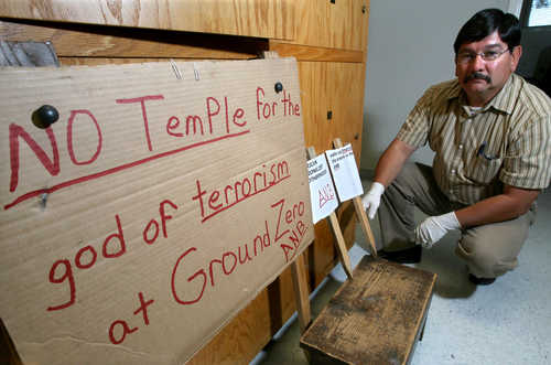 a sad-looking man crouches next to a crudely made cardboard sign that reads No temple for the God of terrorism at Ground Zero