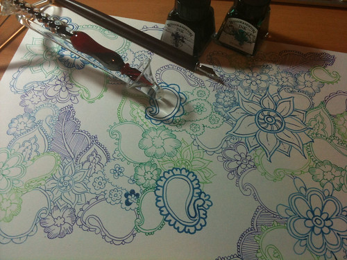 Photo of  a drawing of paisley in different coloured inks (blues and greens). Two small bottles of ink, a glass dip pen and a wooden dip pen with a metal nib sit on top of the paper.