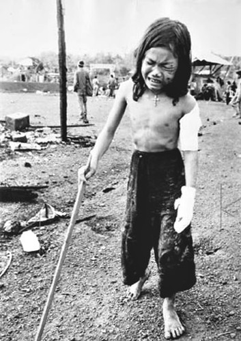 12-yr-old girl with a cross around her neck and w. multiple wounds uses a stick to hobble through debris in Dong Xoai as she attempts to get to an evac helicopter. Her father, sister and a brother were killed and their home destroyed, by Horst Faas 1965