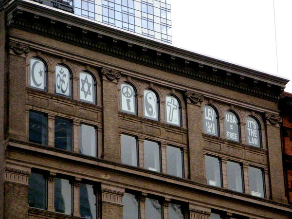 a brick New York apartment building. In each of the top floor windows, a different religious symbol is placed to spell out COEXIST overlooking the Ground Zero site.