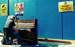 Image of a man playing a tired old piano in front of a building site that has warning signes.  One says, 'danger, men working overhead'.