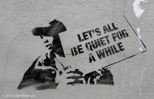 stencil graffiti of a young man holding a sign that reads, let's all be quiet for awhile.