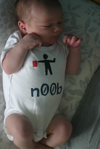 A baby with a white onesie that has a stick figure holding a red semaphore flag, with the word n00b written underneath
