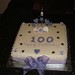 Square 100th birthday cake with lilac/purple heart and star decorations and a champagne bottle.