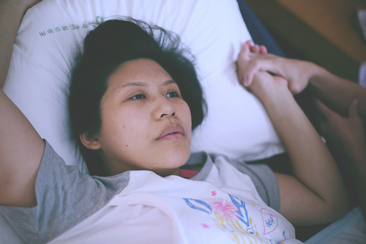孕婦仰躺在枕頭上，一隻手臂抬起，另一隻手與床旁的人相握。