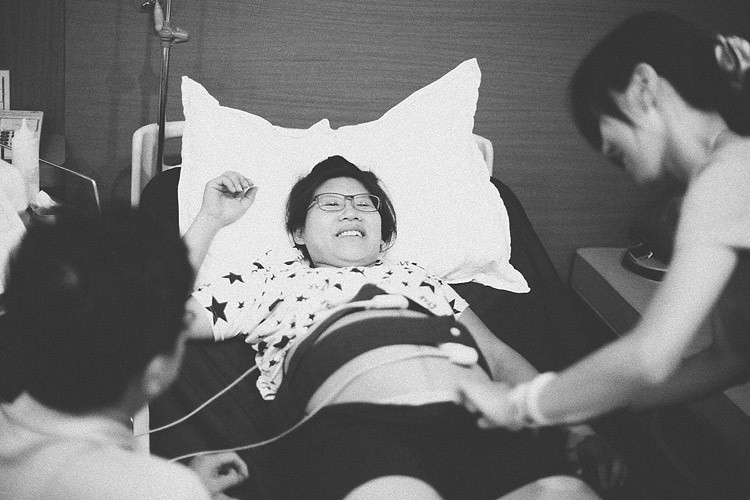孕婦躺在待產床上閉眼抬起手臂，姐姐與助產師分站在床邊。